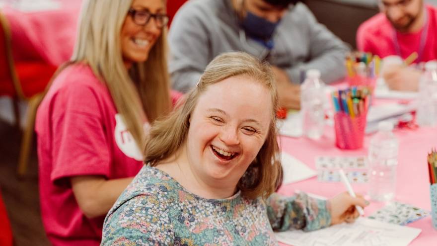 A young woman with downs syndrome smiling at the camera sitting at a round table with other people including Mencap care workers doing arts and crafts