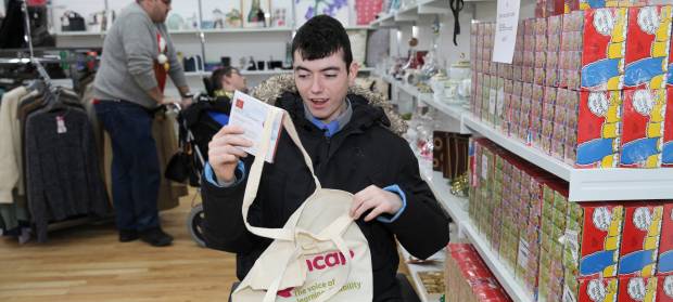 Young man holding Mencap tote bag, browsing items in charity shop.