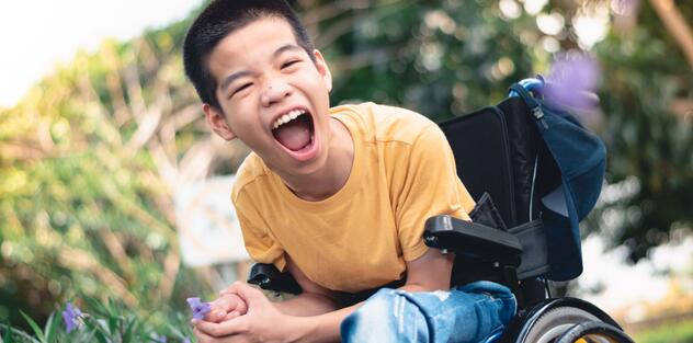 A young boy in a wheelchair who is bending forward with his mouth open shouting happily to the camera