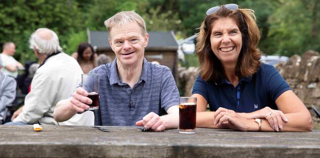 A man with a learning disability with his support worker having a drink of Coke sitting on a wooden picnic bench