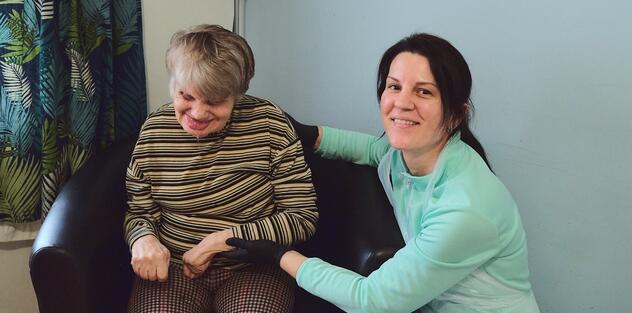 A support worker is crouched down next to the lady she cares for who is sitting on a chair